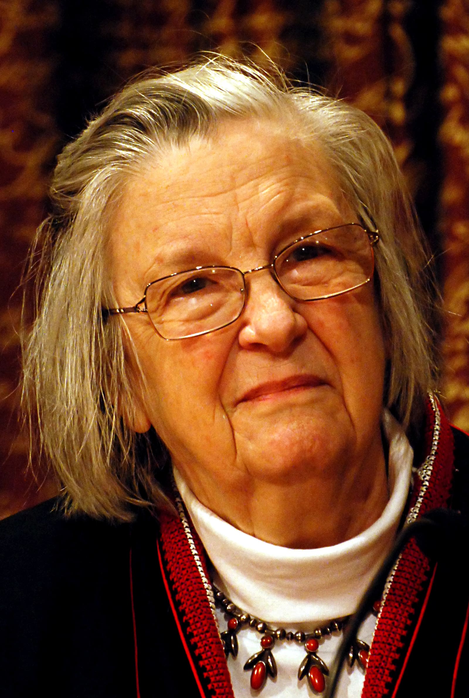 Elinor Ostrom alla conferenza stampa del Nobel a Stoccolma nel dicembre 2009, con capelli grigi e occhiali, dolcevita bianco, giacca scura con bordo rosso e una collana con pietre rosse.