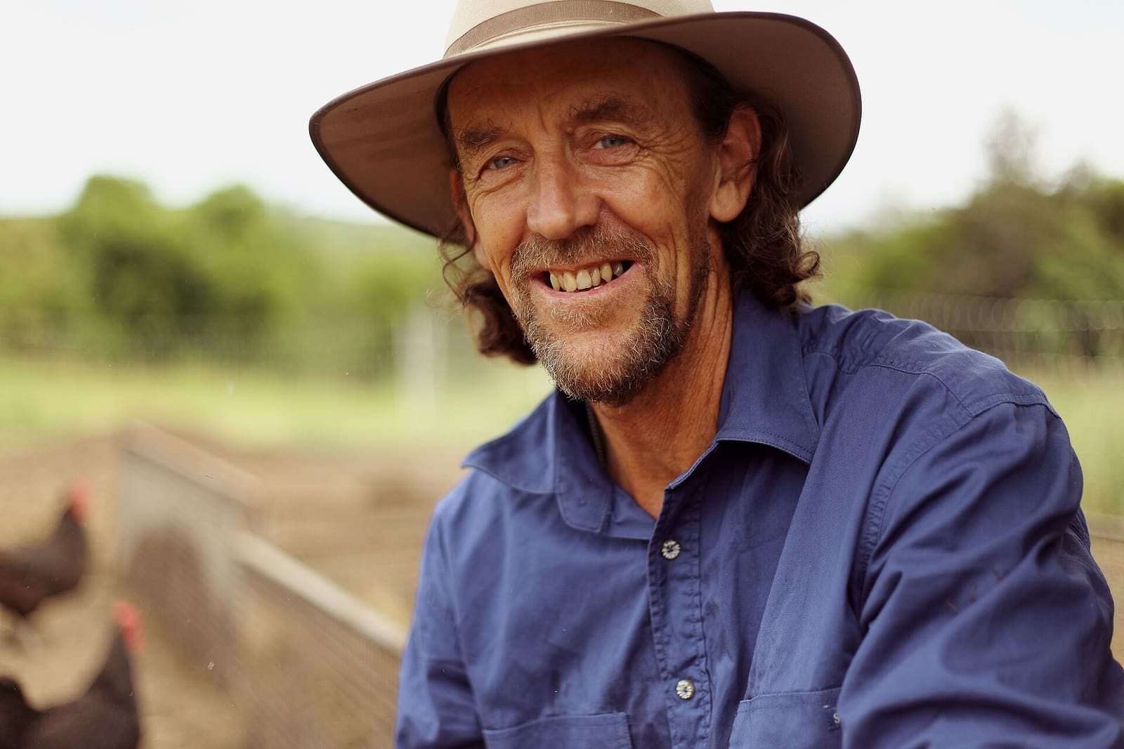 Geoff Lawton in 2015, wearing a wide-brimmed hat and blue shirt, outdoors next to a chicken run.