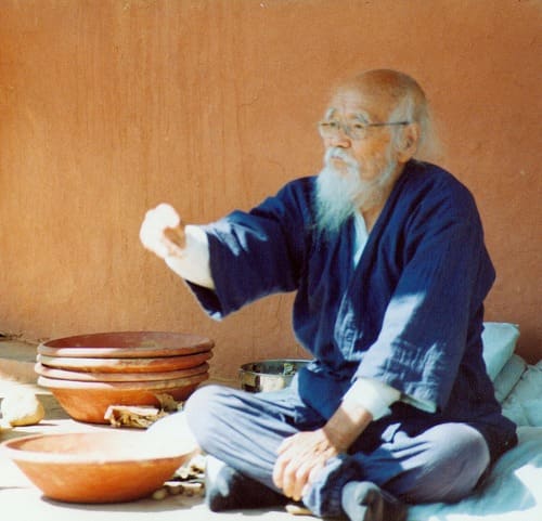 Masanobu Fukuoka sitting cross-legged on the ground, throwing a seed ball, at a workshop at Navdanya, India, in October 2002.