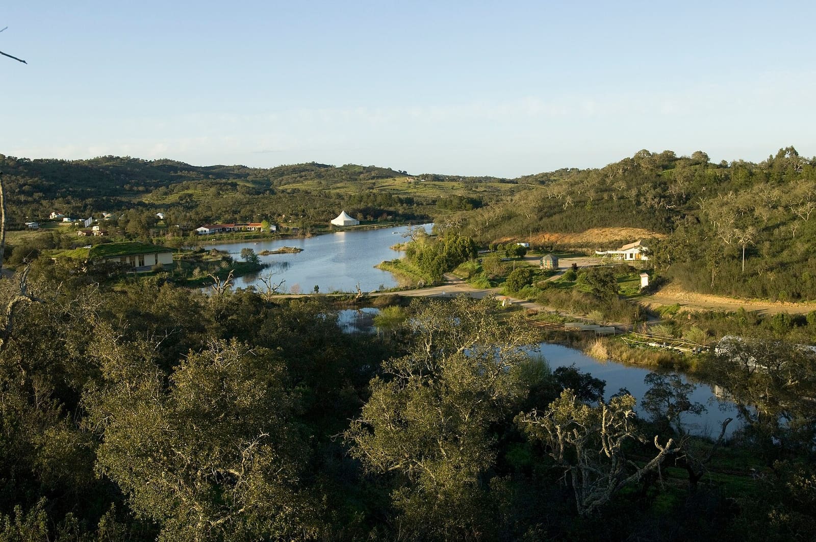 Water retention landscape at Tamera, Portugal: a lake in a wooded valley with buildings along the shore, in evening light.