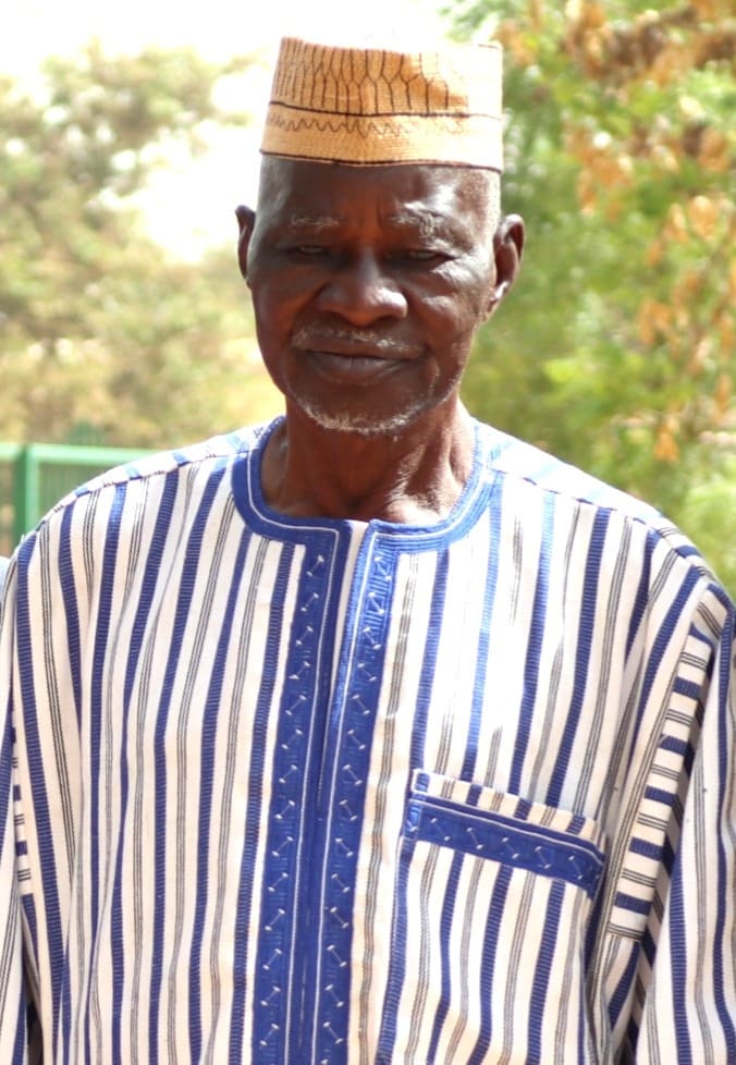 Yacouba Sawadogo in 2019, standing outdoors in a blue and white striped robe and a woven cap.